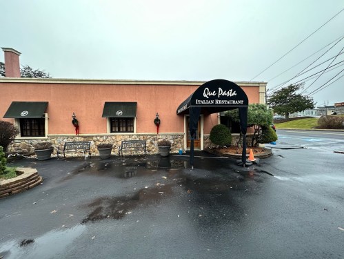 Exterior of Que Pasta Italian Restaurant, featuring a covered entrance and benches, on a rainy day.