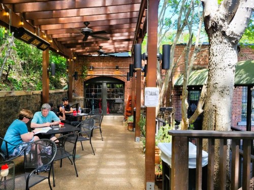 Outdoor dining area with tables, chairs, and greenery, featuring patrons enjoying their meals under a wooden pergola.