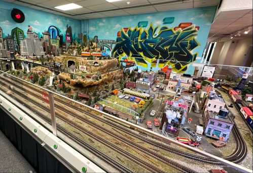 A detailed model train setup featuring a cityscape, mountains, and graffiti art on the wall in the background.