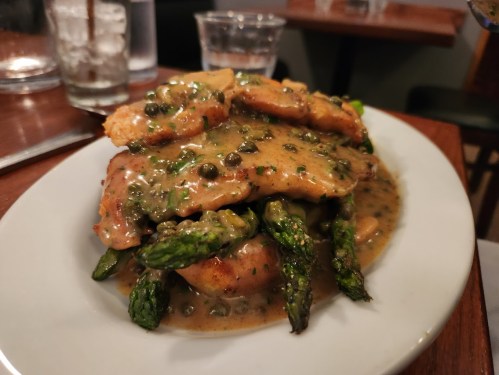 A plate of chicken topped with caper sauce, served over asparagus and garnished with herbs.