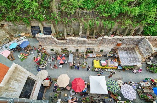 Aerial view of a vibrant outdoor market with colorful umbrellas, people, and rustic buildings surrounded by greenery.