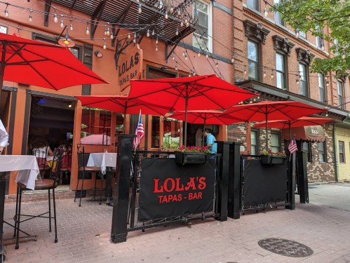Outdoor seating area of Lola's Tapas Bar with red umbrellas and American flags, set against a brick building.