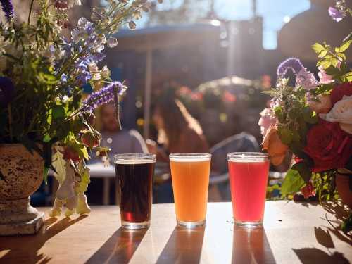 Three glasses of colorful drinks sit on a table, surrounded by vibrant flowers, with people blurred in the background.