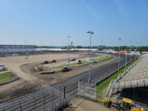 A wide view of a dirt racetrack with maintenance vehicles, grandstands, and clear blue skies in the background.