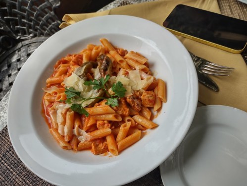 A plate of penne pasta in a creamy tomato sauce, garnished with herbs and cheese, next to a smartphone and fork.