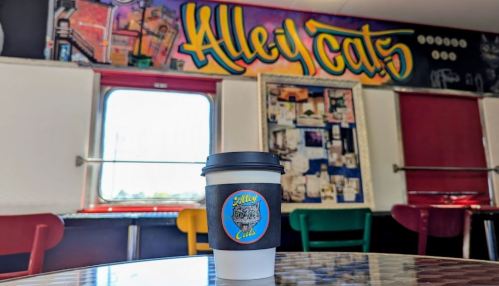 A coffee cup sits on a table in a colorful café with a mural reading "Alley Cats" in the background.