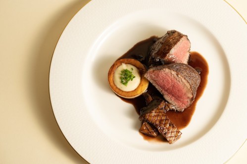 A gourmet plate featuring sliced beef, a potato cake, and garnished with herbs, all drizzled with rich sauce.