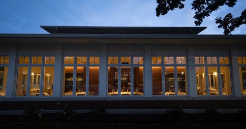 A modern restaurant exterior at dusk, featuring large windows and warm interior lighting.