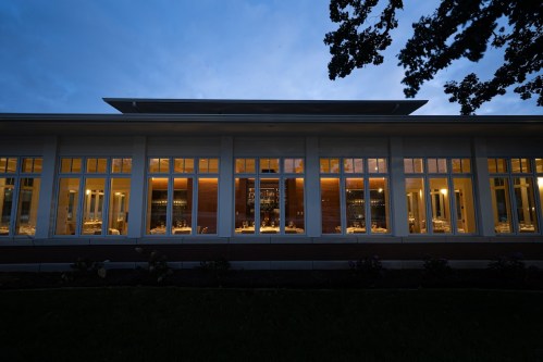 A modern building at dusk, featuring large windows illuminated from within, surrounded by greenery.