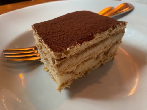 A slice of tiramisu on a white plate, with a fork beside it, showcasing layers of coffee-soaked cake and creamy filling.