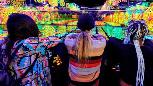 Three people with colorful outfits admire a vibrant, mirrored light installation filled with swirling patterns.