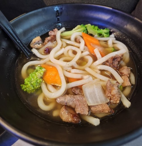 A bowl of udon soup with beef, carrots, broccoli, and onions in a savory broth.