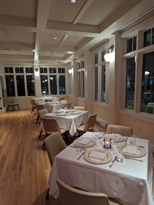 A cozy restaurant interior with elegantly set tables, soft lighting, and large windows.