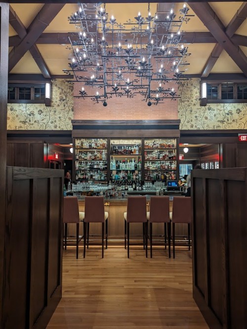 A cozy bar area with a stylish chandelier, wooden beams, and a well-stocked shelf of liquor in a warm, inviting setting.