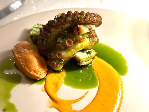 Grilled octopus on a plate with vibrant sauces and a scoop of orange puree, garnished with green herbs.
