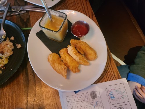 A plate with three chicken tenders, a small cup of dipping sauce, and a side of applesauce on a wooden table.