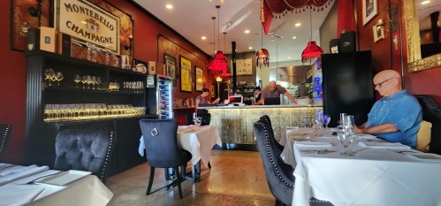 A cozy restaurant interior with red decor, a bar, and a man sitting alone at a table. Elegant table settings are visible.