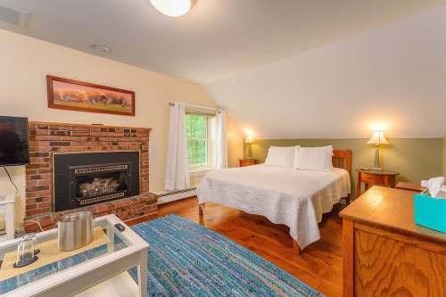 Cozy bedroom with a brick fireplace, a double bed, wooden furniture, and a colorful rug. Natural light from a window.