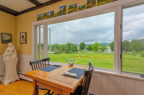 A cozy dining area with a wooden table and chairs, featuring a large window overlooking a green landscape and cloudy sky.