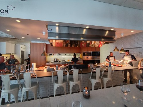 A modern kitchen with chefs preparing food, featuring a large counter and cooking equipment in a bright, open space.