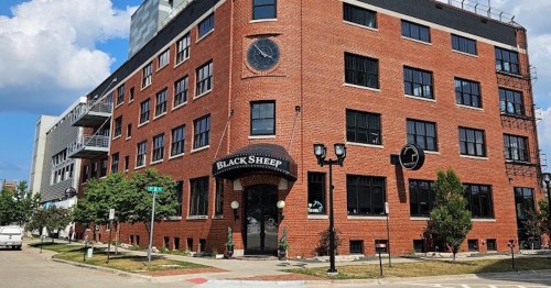 A brick building with large windows and a sign reading "Black Sheep," surrounded by trees and parked cars.