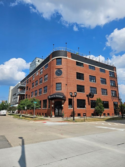 A brick building with large windows and a clock, featuring a sign for "Black Sheep," under a blue sky with clouds.