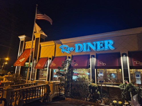 A brightly lit diner at night, featuring a blue neon sign that reads "Red Diner" and an American flag.