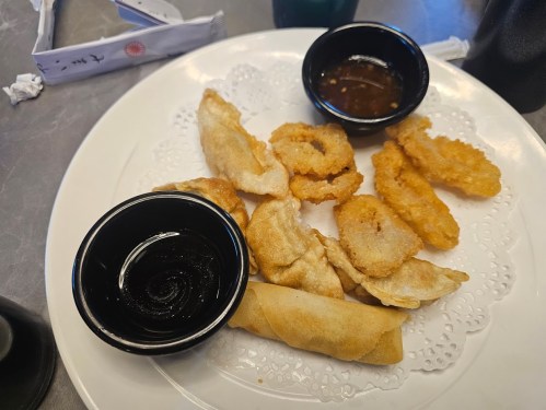 A plate of assorted fried appetizers, including spring rolls and dipping sauces, on a lace doily.