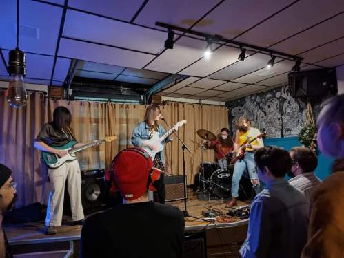A band performs on stage with guitars and drums, while an audience watches in a cozy venue.