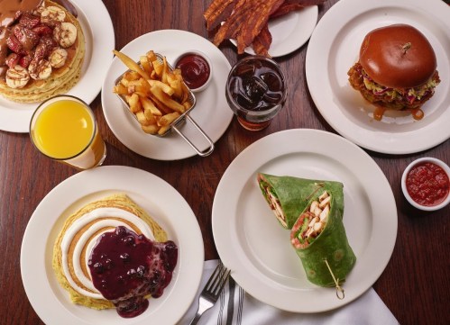 A table spread featuring pancakes with syrup, a wrap, fries, a burger, bacon, and drinks.