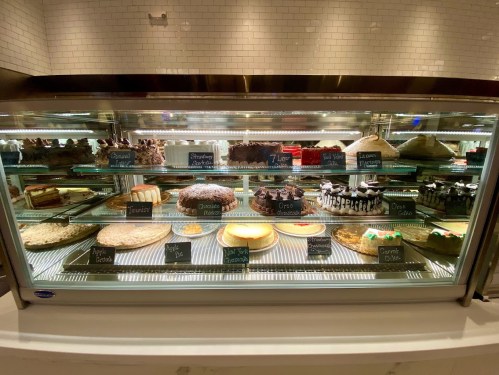 A display case filled with various cakes and desserts, each labeled with names on small chalkboard signs.