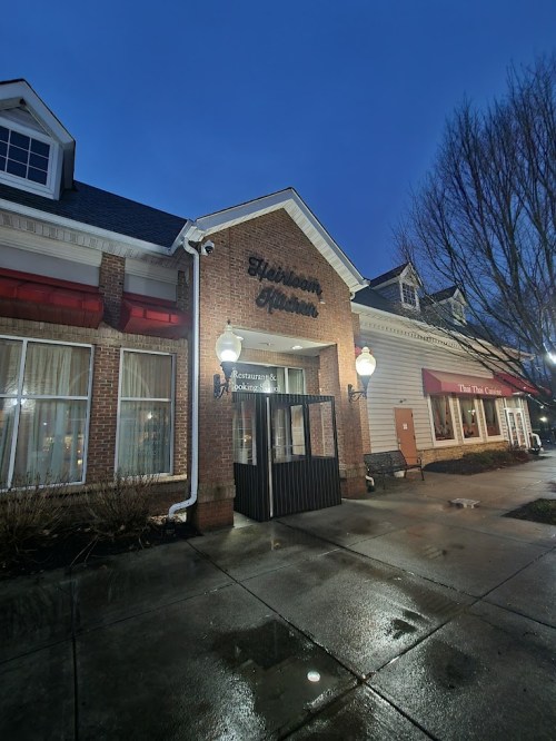 Exterior of a restaurant at dusk, featuring a brick facade, large windows, and illuminated signage. Wet pavement reflects light.