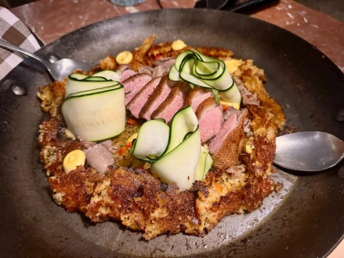 A savory dish featuring sliced meat, cucumber ribbons, and a crispy base, served in a round metal pan.