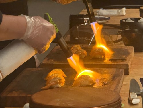A chef uses a torch to sear sushi on wooden boards, creating flames above the delicacies.