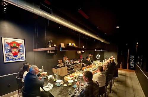 A group of people dining at a sushi restaurant, seated at a long table with various dishes and a chef preparing food.