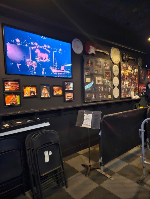 A dimly lit venue wall featuring a TV screen, framed photos, and musical memorabilia. Chairs are stacked nearby.