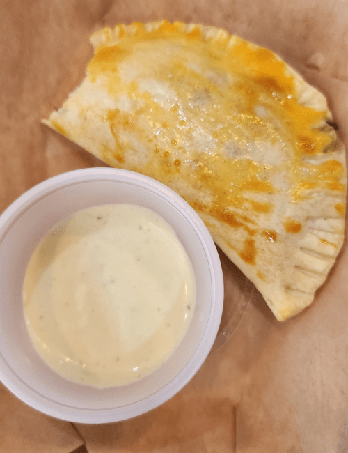A golden-brown calzone on parchment paper, accompanied by a small cup of creamy dipping sauce.