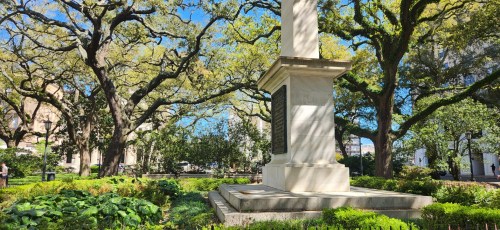 A stone monument surrounded by lush greenery and large trees in a sunny park setting.