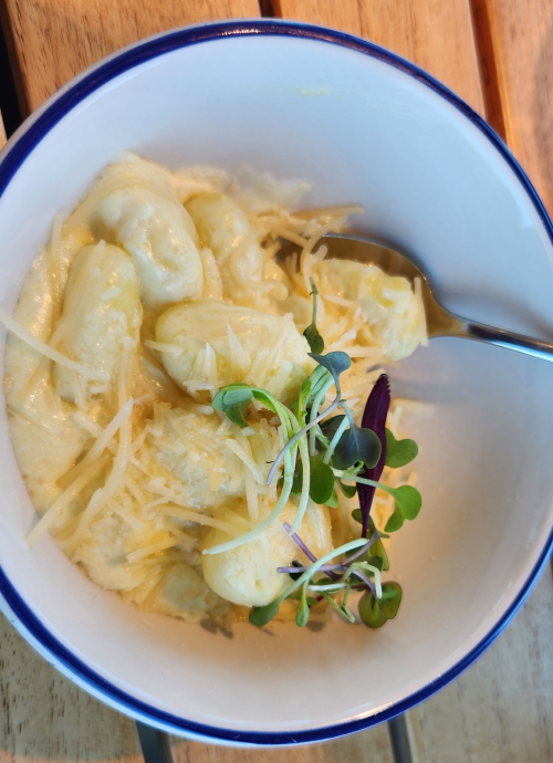 A bowl of creamy gnocchi topped with grated cheese and microgreens, served with a spoon on a wooden table.