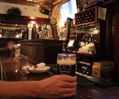 A hand holding a pint of Guinness at a bar, with bottles and taps in the background. Soft lighting creates a cozy atmosphere.