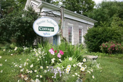 A stone cottage with a sign reading "Stone School Cottage," surrounded by colorful flowers and greenery.