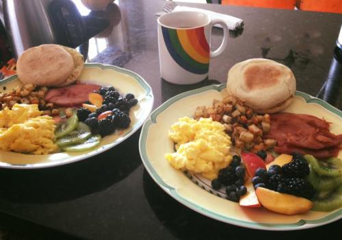 Two plates of breakfast with scrambled eggs, ham, potatoes, fruit, and a rainbow mug on a black countertop.