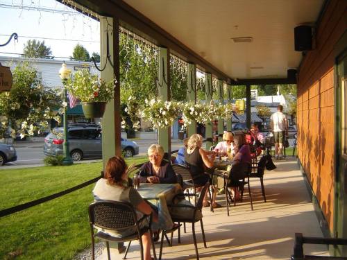 A cozy outdoor café scene with people dining at tables, surrounded by hanging flower baskets and warm evening light.