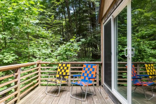 Two colorful chairs on a wooden deck surrounded by lush green trees. A large glass door reflects the scenery.