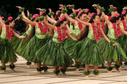 A group of dancers in green outfits and floral leis perform a traditional hula dance on a stage.