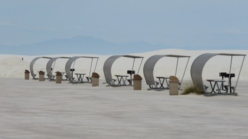 A row of shaded picnic tables on a flat, sandy landscape with distant mountains under a cloudy sky.