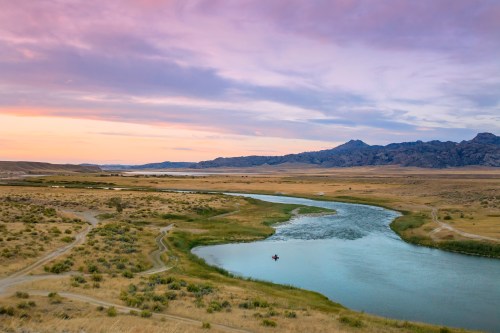 A serene river winding through golden grasslands under a colorful sunset, with mountains in the background.