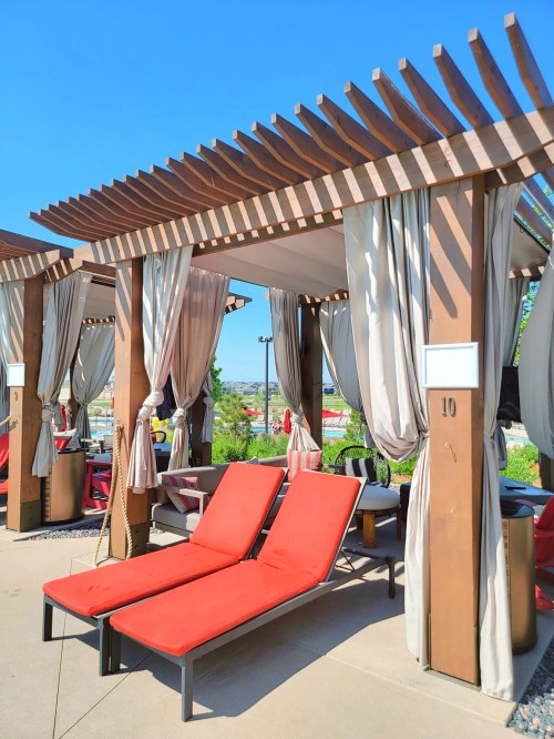 Two red lounge chairs under a wooden pergola with draped curtains, set against a clear blue sky.