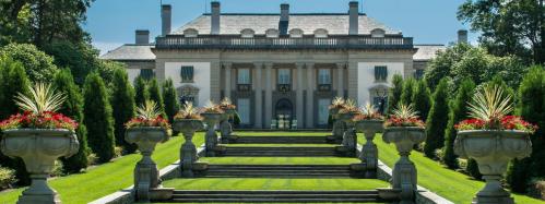 A grand mansion with a symmetrical facade, surrounded by lush greenery and flower-filled planters along a staircase.