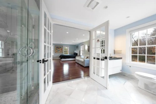 Bright bathroom with glass shower, open double doors leading to a spacious bedroom with a bed and seating area.